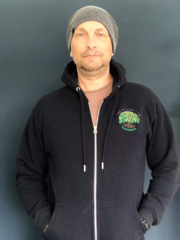A man in a grey beanie and black hoodie with a "Willow Tree Foundation" logo looks calmly at the camera.