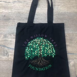 Black tote bag featuring an embroidered green weeping willow tree logo and "THE WILLOW TREE FOUNDATION" text on a wooden floor.