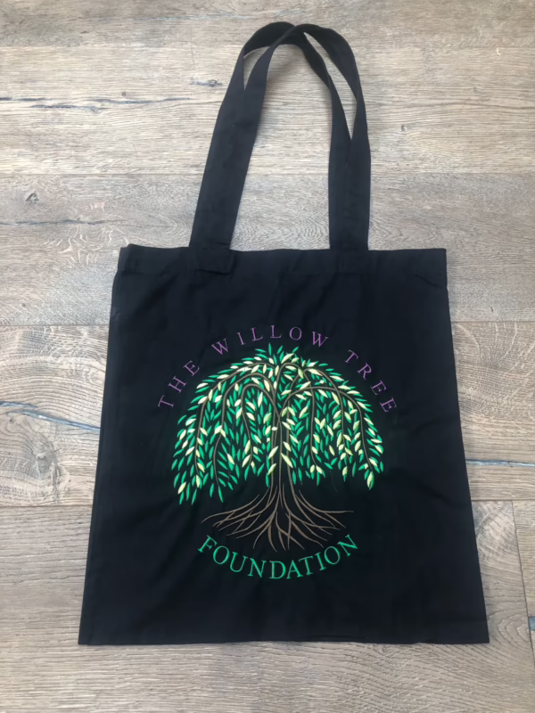 Black tote bag featuring an embroidered green weeping willow tree logo and "THE WILLOW TREE FOUNDATION" text on a wooden floor.