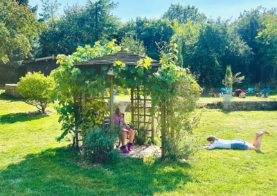 Two people relax in a sunny, lush garden, one reading in a vine-covered gazebo and another lying on the green grass.