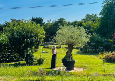 Woman stands beside an olive tree in a lush, sunny garden where another person relaxes on the grass.