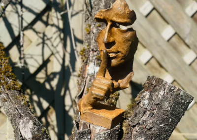 Rustic golden sculpture of a face with a finger on its lips, placed on a mossy tree stump against a sun-dappled wooden fence.